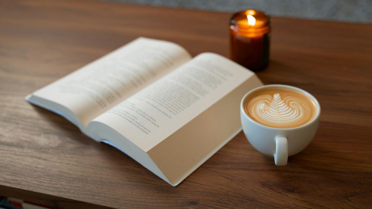 An open book, a latte with latte art, and a lit amber jar candle on a wooden table