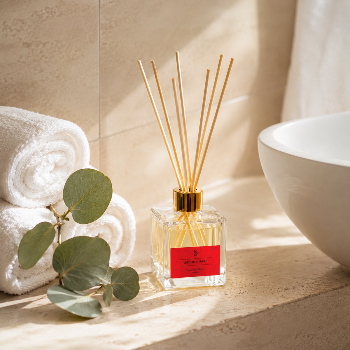 The Amour a Paris reed diffuser on a bathroom ledge with rolled towels, eucalyptus and a basin