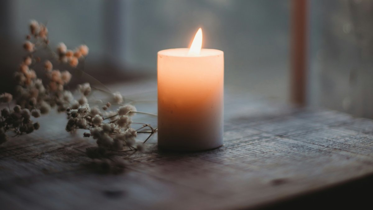 A lit pillar candle on a wooden windowsill at dusk with dried flowers and a winter view beyond the window