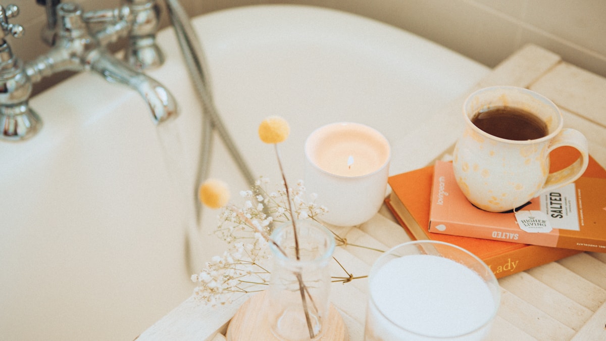 A lit candle, a cup of tea, a book and dried flowers on a wooden tray across a bath