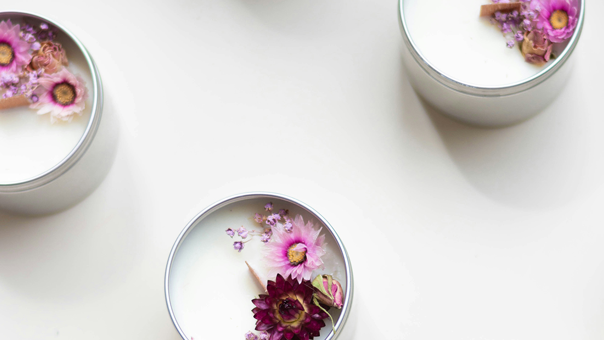 Three soy candle tins topped with dried pink and purple flowers on a white surface